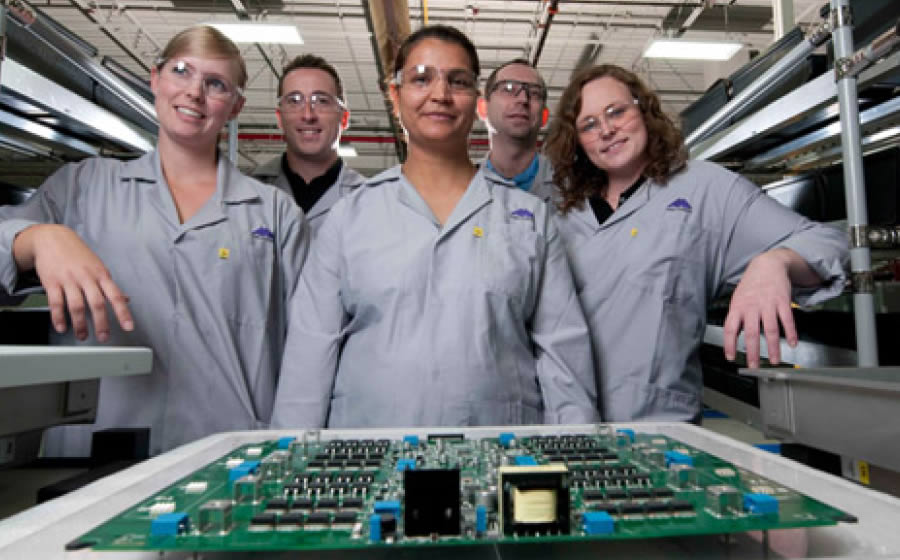 Five Melitron employees with safety glasses pose in front of a circuit board in Melitron's manufacturing facility.